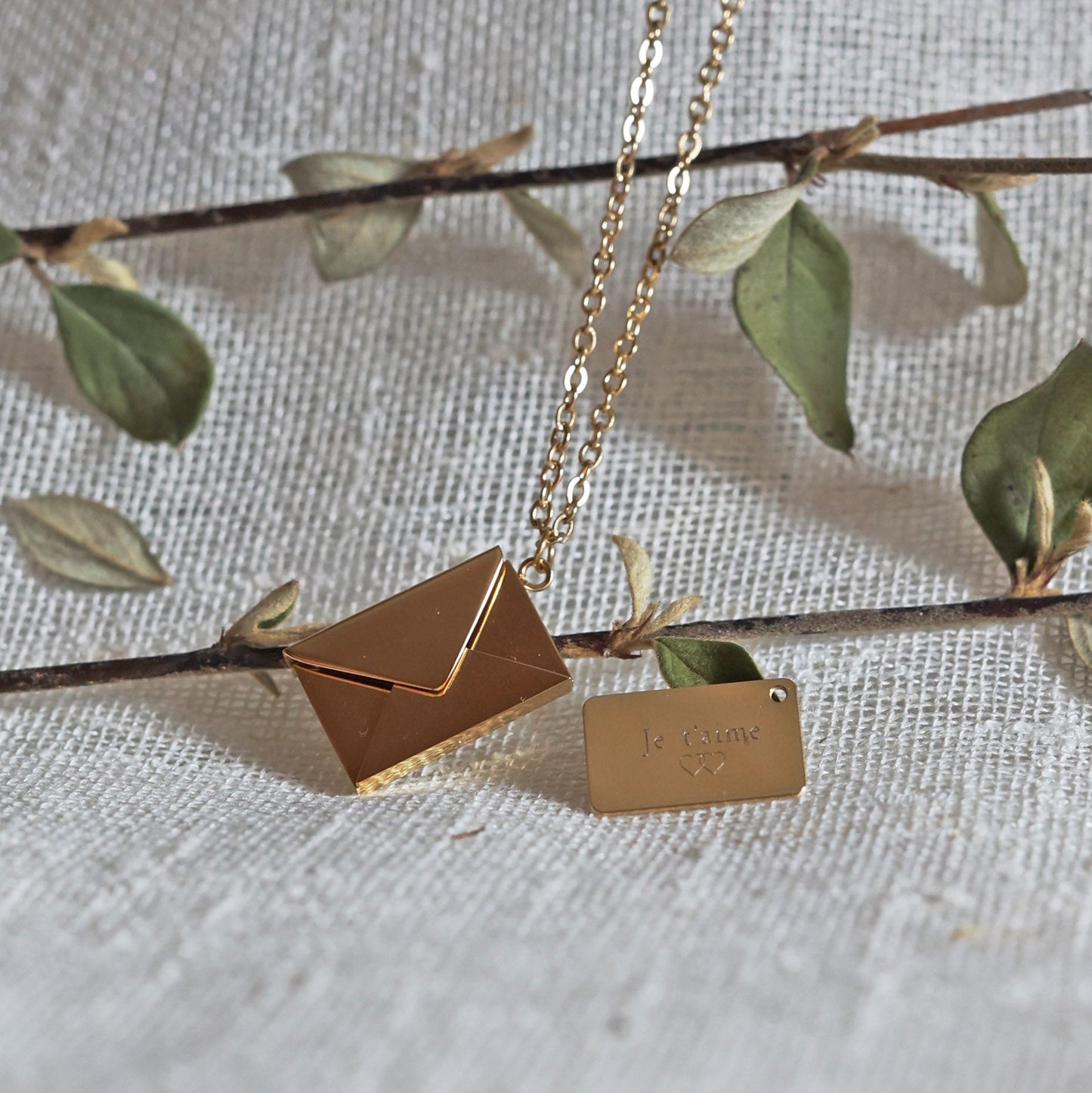 Collier "Nid secret", pendentif en forme d'enveloppe dorée ouverte, dévoilant un message "je t'aime", posé avec soin sur un fond beige texturé et sur une branche de feuilles