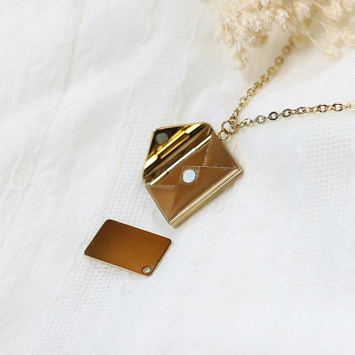 Collier "Nid secret", pendentif en forme d'enveloppe dorée ouverte, posé avec soin sur un fond beige texturé.