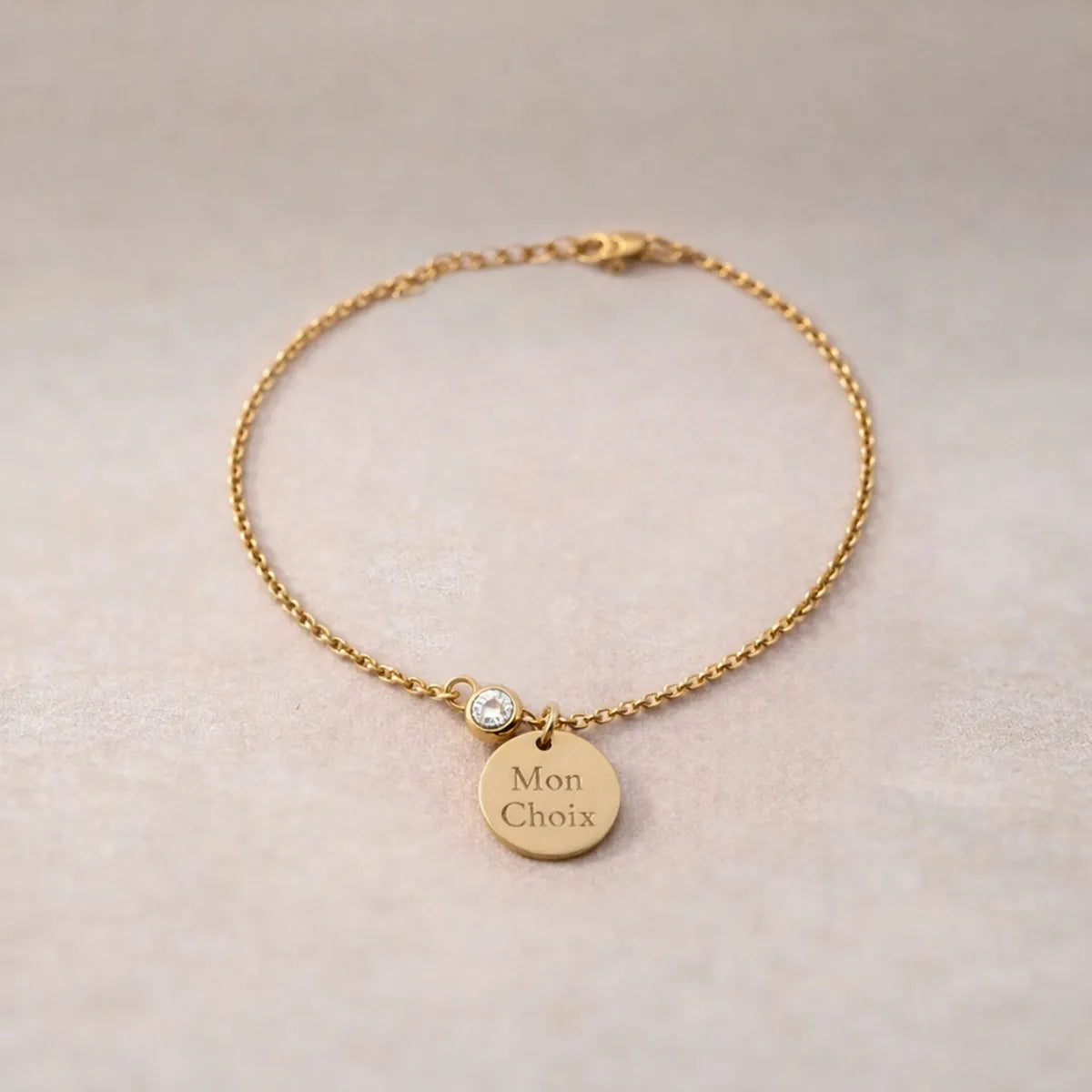 Bracelet Mon choix doré, bijou personnalisable en acier inoxydable avec médaillon gravé "Mon choix" et couleur de naissance blanche, posé sur un fond beige texturé.
