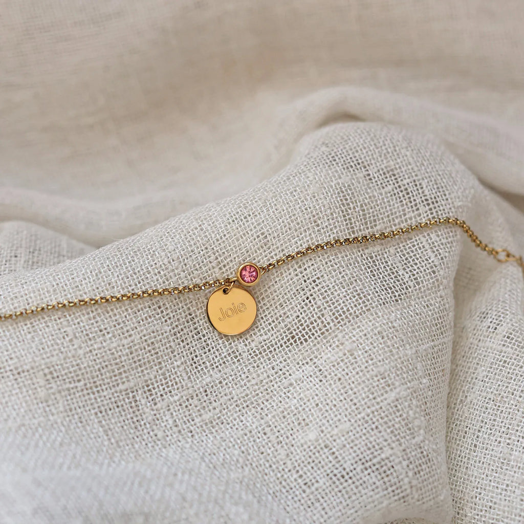 Bracelet Mon choix doré, bijou personnalisable en acier inoxydable avec médaillon gravé "Joie" et pierre de naissance rose, posé sur un tissu beige