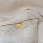 Bracelet Mon choix doré, bijou personnalisable en acier inoxydable avec médaillon gravé "Joie" et pierre de naissance rose, posé sur un tissu beige