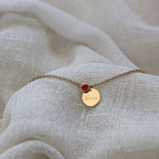 Bracelet Mon choix doré, bijou personnalisable en acier inoxydable avec médaillon gravé "Amour" et pierre de naissance rouge, posé sur un tissu beige
