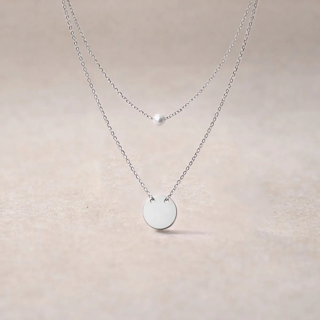 Collier Visée Complice double chaîne perle et médaille acier inoxydable argenté, sur fond beige texturé