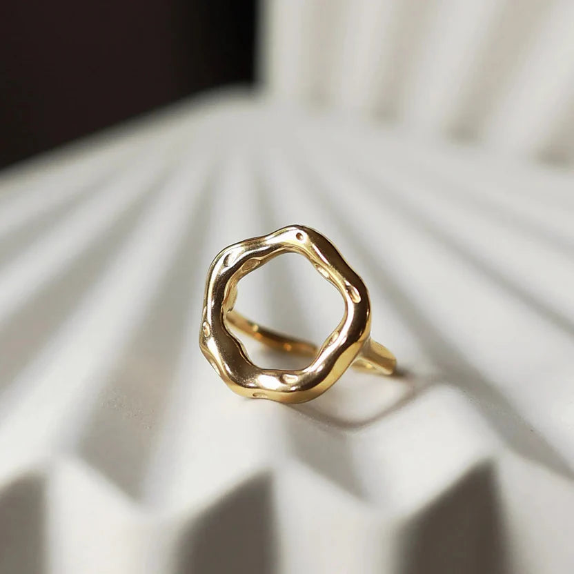 Bague Mon tout, au design cercle martelé en acier inoxydable doré, posée sur un décor beige texturée.