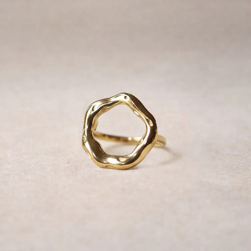 Bague Mon tout, au design cercle martelé en acier inoxydable doré, posée sur un décor beige texturée.