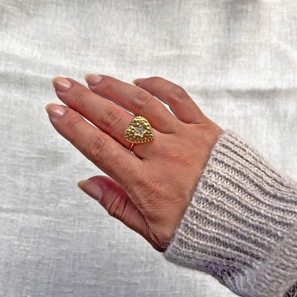 Bague ajustable Coeur battant, en acier doré en forme de cœur finement martelé, ornée d'une étoile brillante au centre, portée à la main sur fond beige. 
