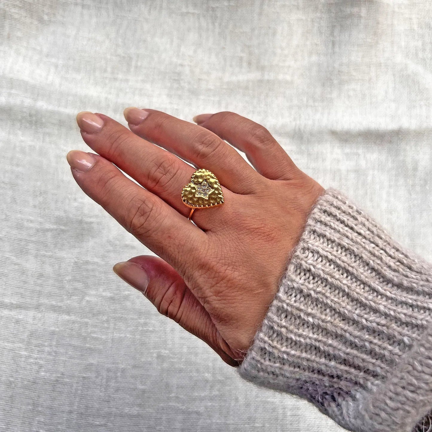 Bague ajustable Coeur battant, en acier doré en forme de cœur finement martelé, ornée d'une étoile brillante au centre, portée à la main sur fond beige. 