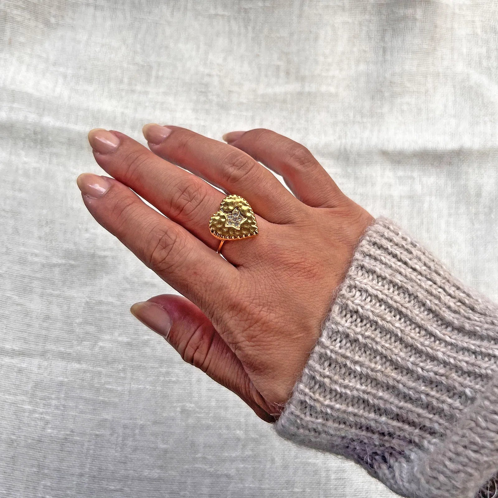 Bague ajustable Coeur battant, en acier doré en forme de cœur finement martelé, ornée d'une étoile brillante au centre, portée à la main sur fond beige. 