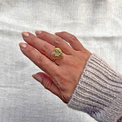 Bague ajustable Coeur battant, en acier doré en forme de cœur finement martelé, ornée d'une étoile brillante au centre, portée à la main sur fond beige. 