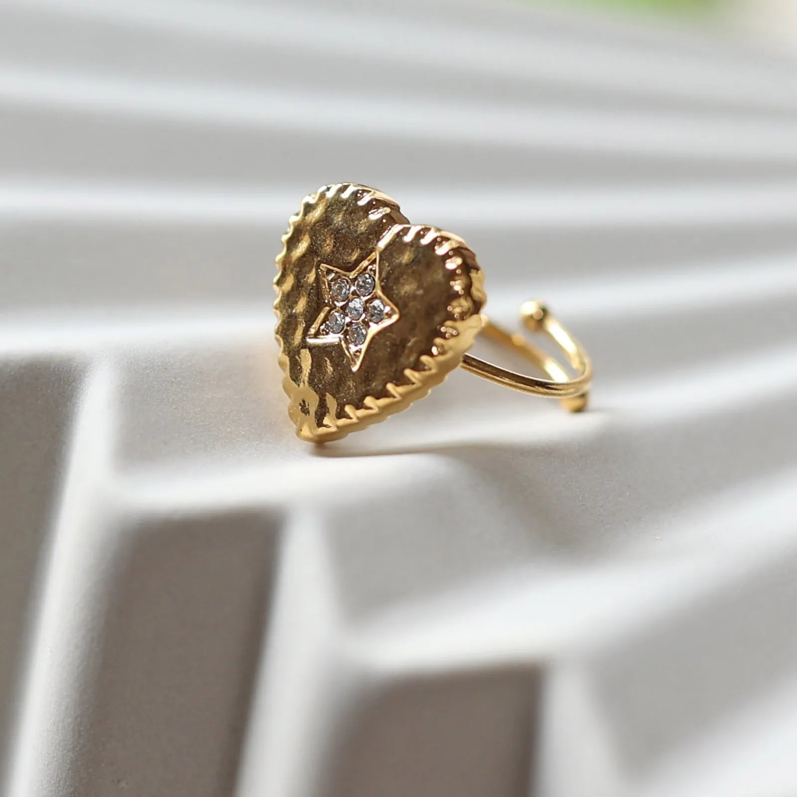 Bague ajustable Coeur battant, en acier doré en forme de cœur finement martelé, ornée d'une étoile brillante au centre, photographiée de profil sur un fond beige texturé.