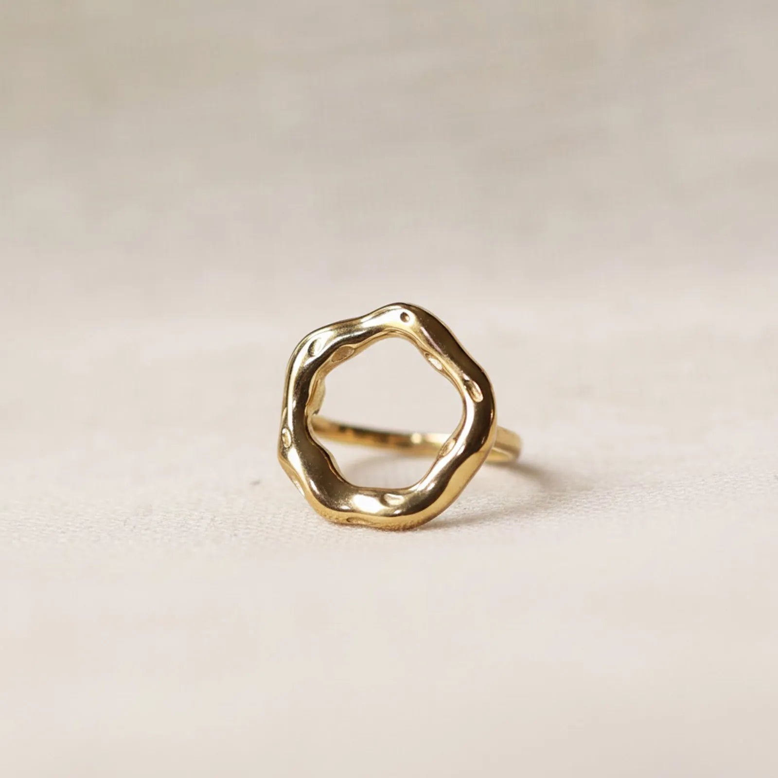 Bague Mon tout, au design cercle martelé en acier inoxydable doré, posée sur un décor beige texturée.