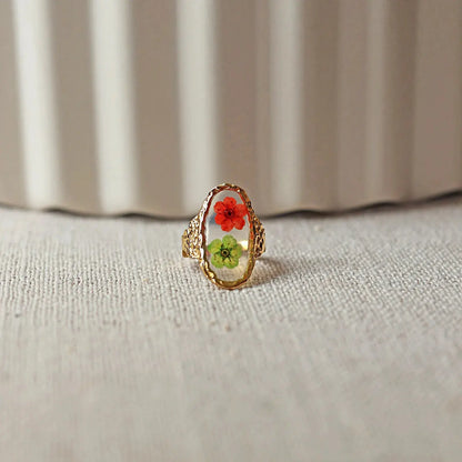 Bague ajustable Mon rythme,en acier inoxydable doré, ornée de deux fleurs séchées rouge et verte, posée à plat sur un tissu beige texturé, vue de face.