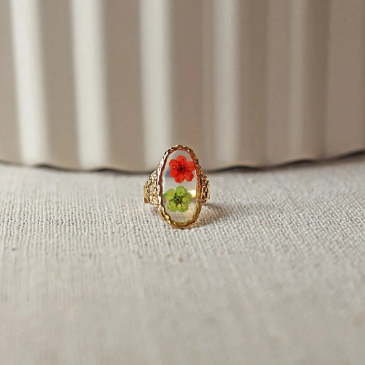 Bague ajustable Mon rythme,en acier inoxydable doré, ornée de deux fleurs séchées rouge et verte, posée à plat sur un tissu beige texturé, vue de face.