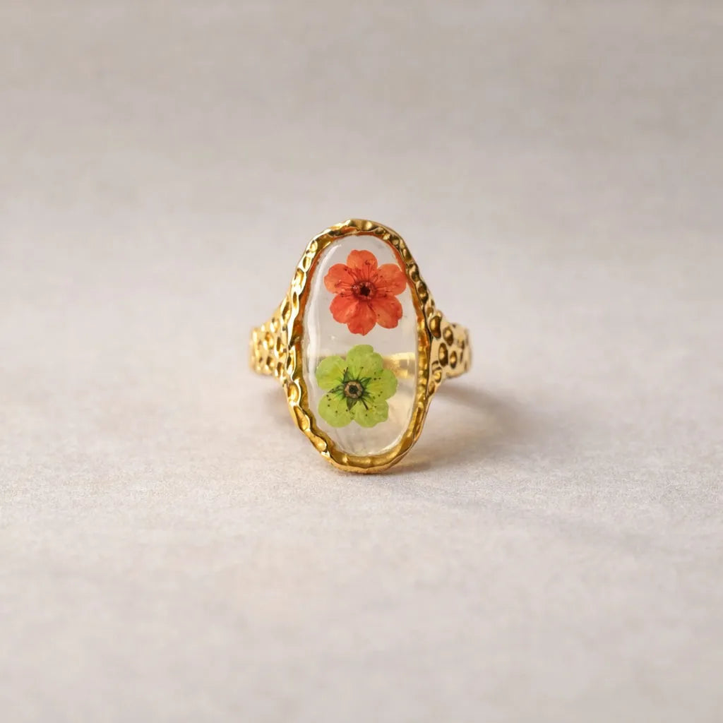 Bague ajustable Mon rythme,en acier inoxydable doré, ornée de deux fleurs séchées rouge et verte, posée à plat sur un tissu beige texturé, vue de face.