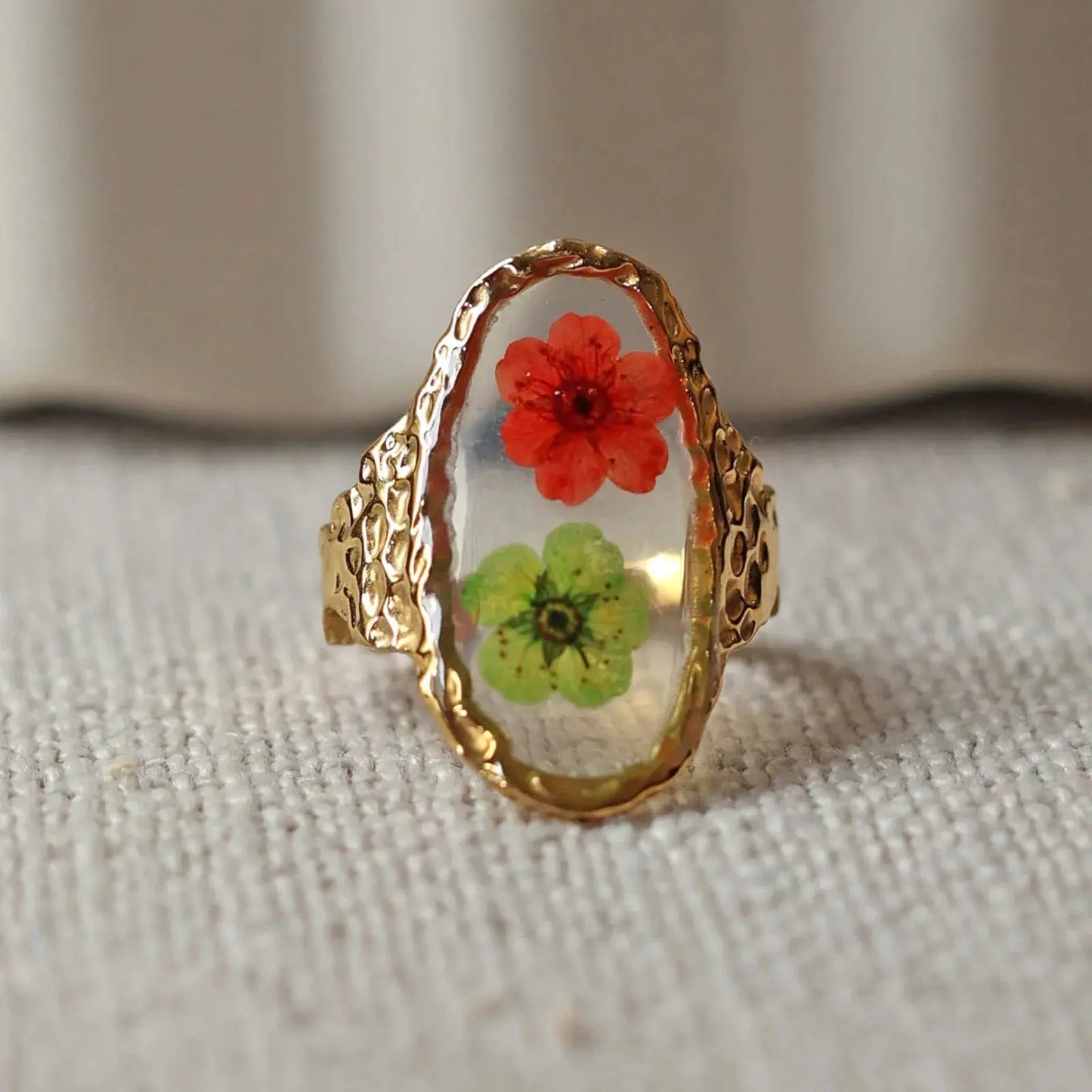 Bague ajustable Mon rythme, en acier inoxydable doré, orné de deux fleurs rouge et verte, posée à plat sur un tissu beige texturé, vue de face rapprochée.