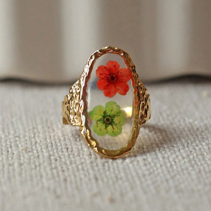 Bague ajustable Mon rythme, en acier inoxydable doré, orné de deux fleurs rouge et verte, posée à plat sur un tissu beige texturé, vue de face rapprochée.
