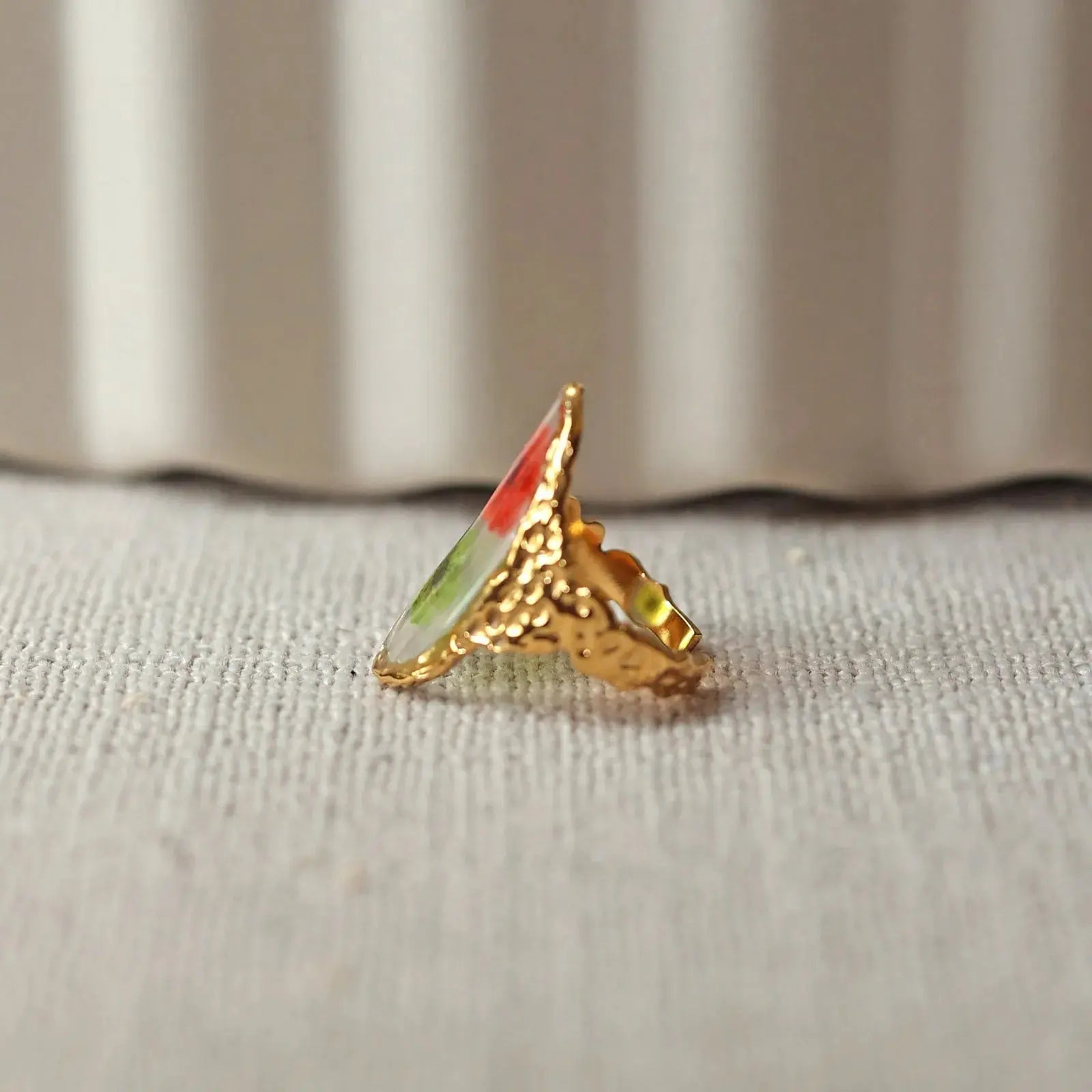 Bague ajustable Mon rythme, en acier inoxydable doré, orné de deux fleurs rouge et verte, posée à plat sur un tissu beige texturé, vue de profil rapprochée.