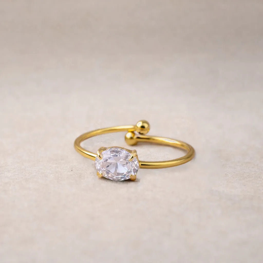 Bague Facette ajustable, en acier doré, ornées d'une pierre de couleur blanche, posées à plat sur un fond beige texturé.