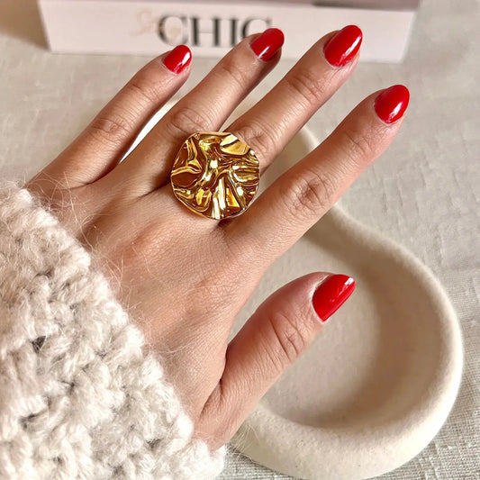 Bague réglable en acier inoxydable doré à motif sculpté, portée sur une main féminine avec vernis rouge, fond beige doux et boîte chic en arrière-plan.