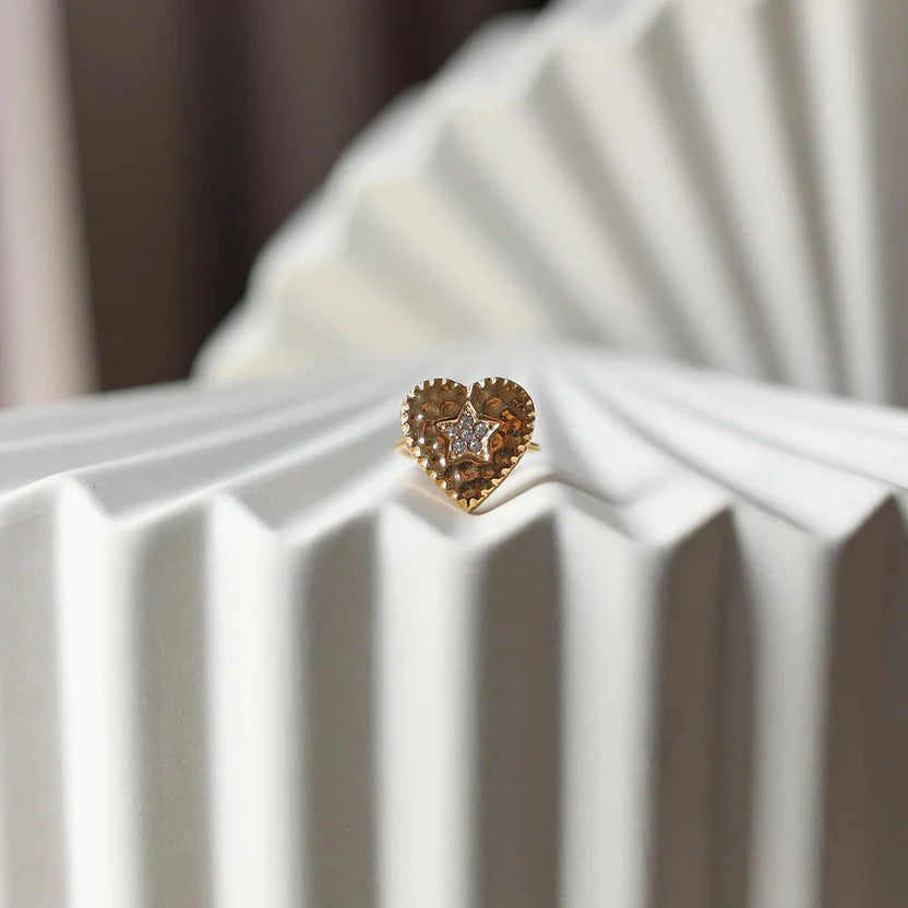 Bague ajustable Coeur battant, en acier doré en forme de cœur finement martelé, ornée d'une étoile brillante au centre, photographiée de face sur un fond beige texturé.