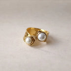 Bague Rencontre ouverte en acier inoxydable doré, ornée de deux perles blanches , vue posée sur un support texturé beige clair.
