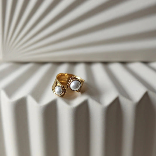 Bague Rencontre ouverte en acier inoxydable doré, ornée de deux perles blanches , vue posée sur un support texturé beige clair.