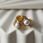 Gros plan sur la bague Rencontre ouverte en acier inoxydable doré, ornée de deux perles blanches, posée sur un support texturé beige clair.