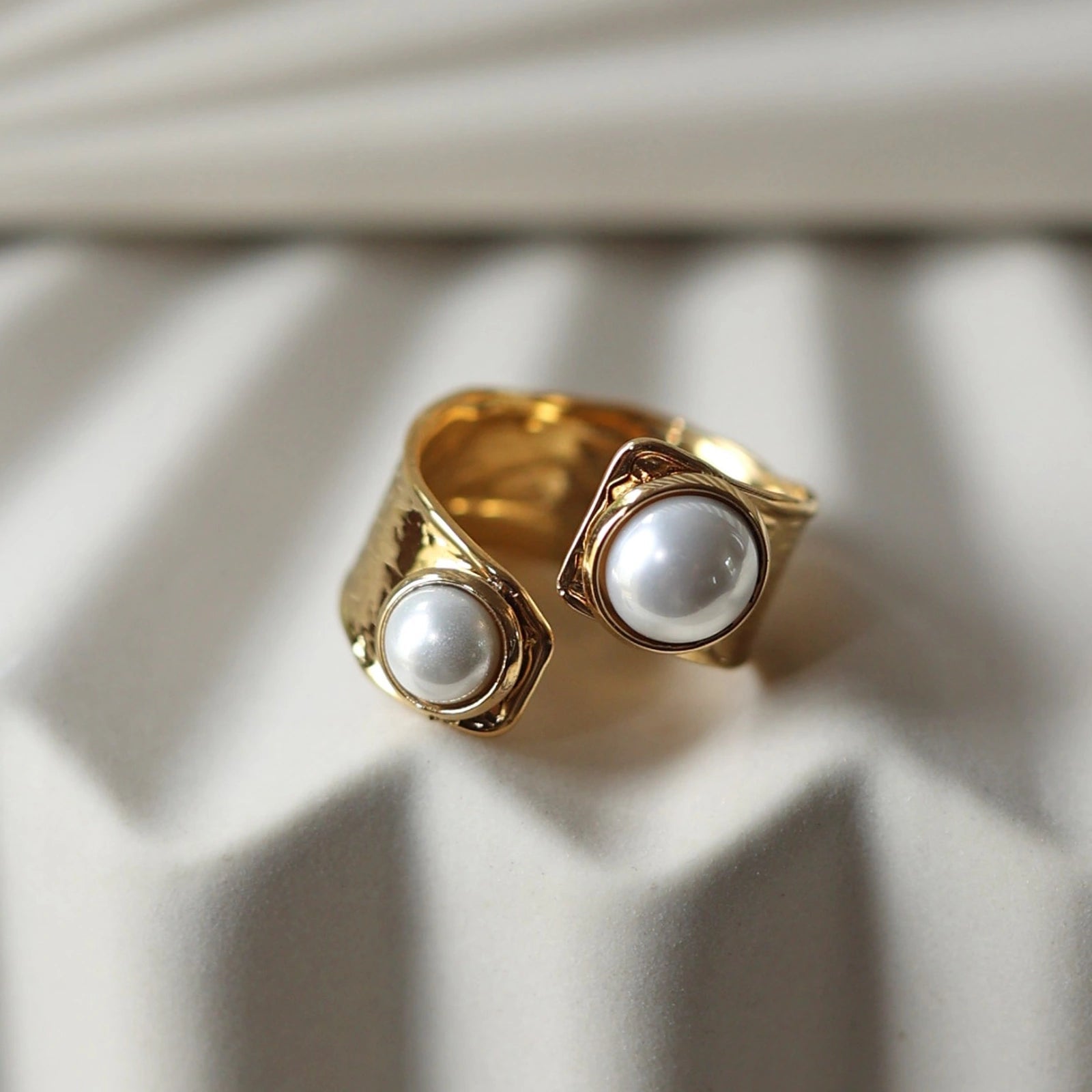 Gros plan sur la bague Rencontre ouverte en acier inoxydable doré, ornée de deux perles blanches, posée sur un support texturé beige clair.