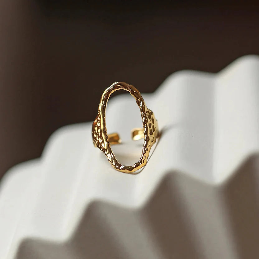 Bague Maintenant en acier inoxydable doré, ovale martelée et réglable, présentée sur un fond beige texturé.