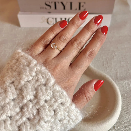 Bague "Mes Facettes" ajustable, en acier doré, ornée d'une pierre blanche, portée sur une main féminine au vernis rouge. En arrière-plan, un livre au style inspiré.