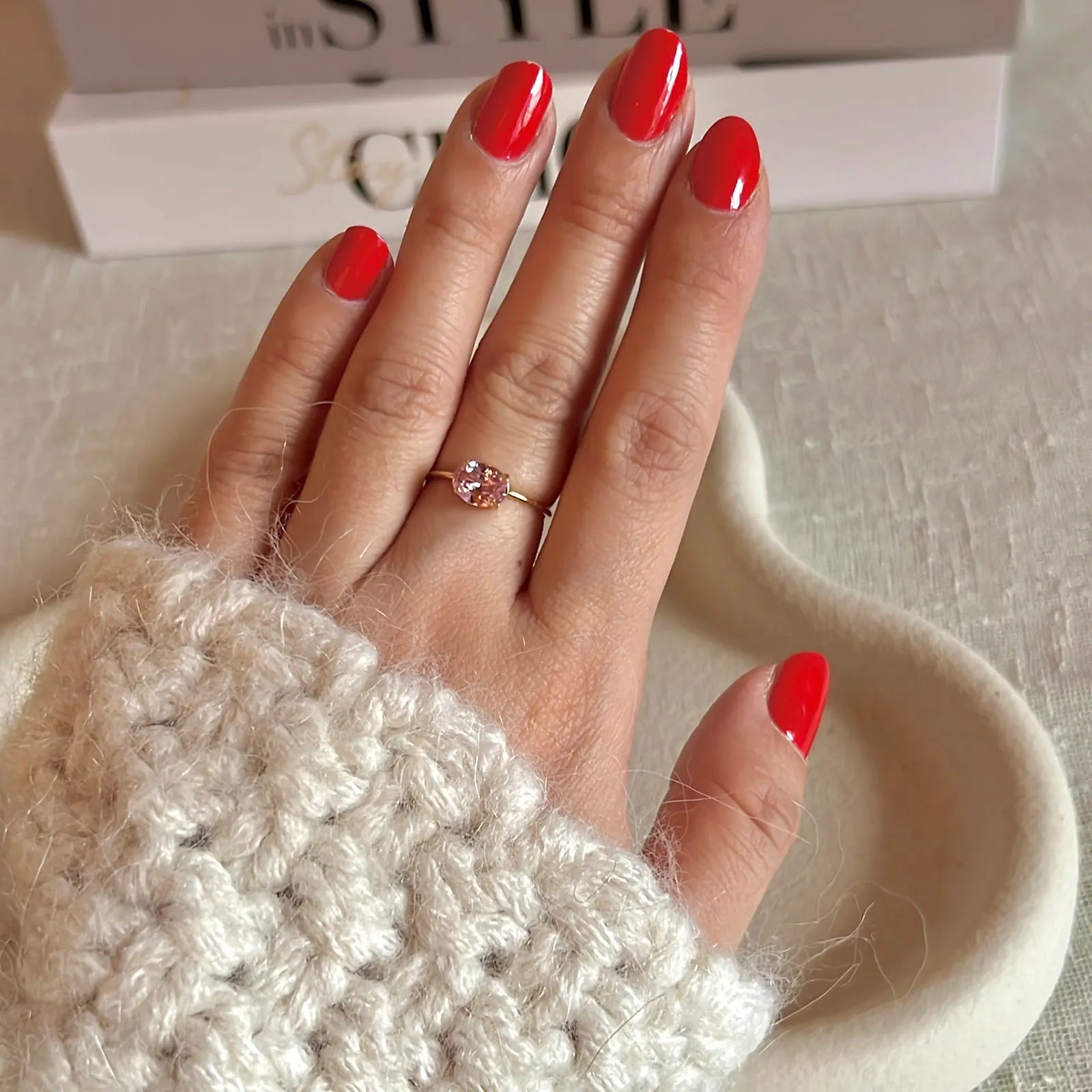 Bague "Mes Facettes" ajustable, ornée d'une pierre rose, portée sur une main féminine au vernis rouge. En arrière-plan, un livre au style inspiré.