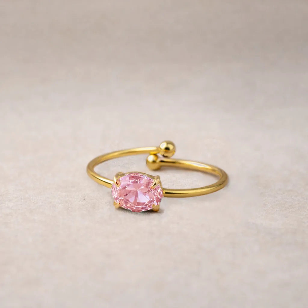 Bagues "Mes Facettes" ajustables, en acier doré, ornées d'une pierre de couleur rose, posées à plat sur un fond beige texturé.