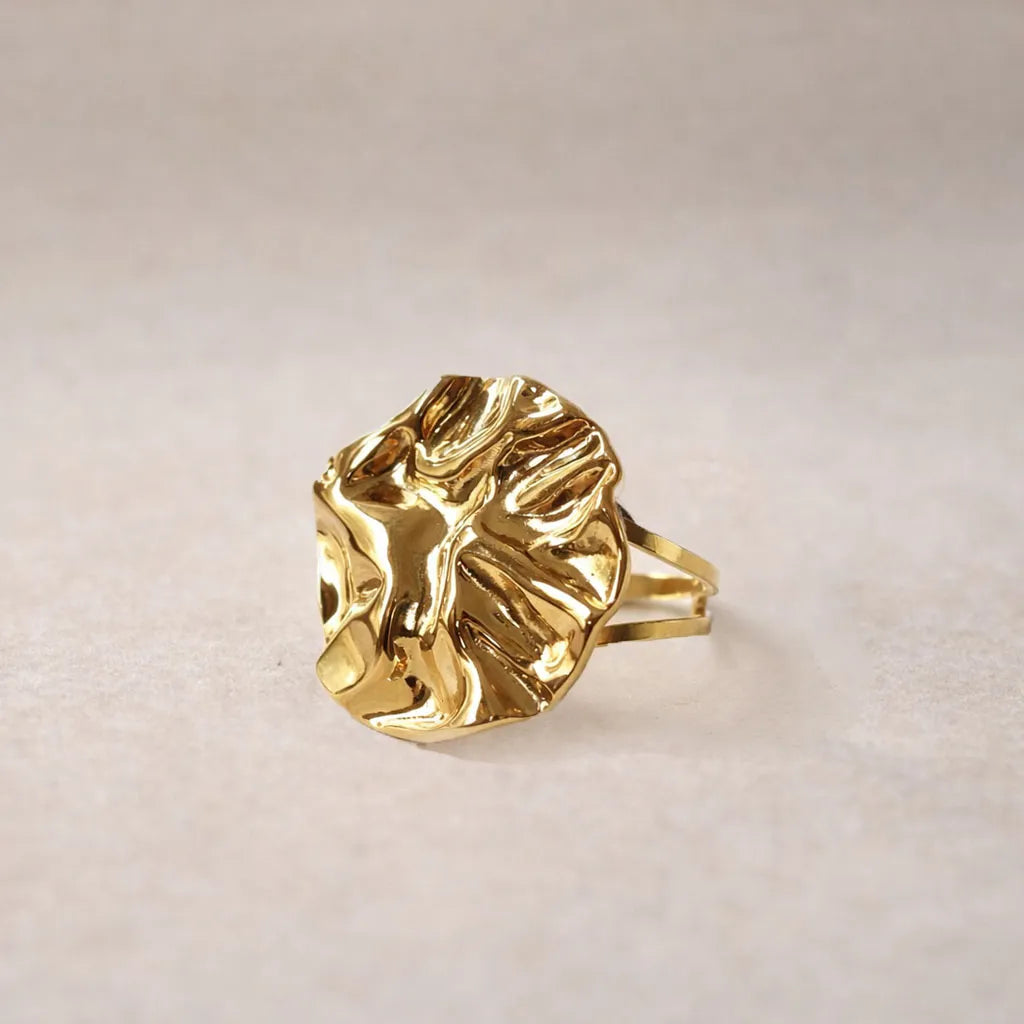 Zoom sur la bague Froissé, réglable en acier inoxydable doré à motif sculpté, posée sur un fond beige texturé.