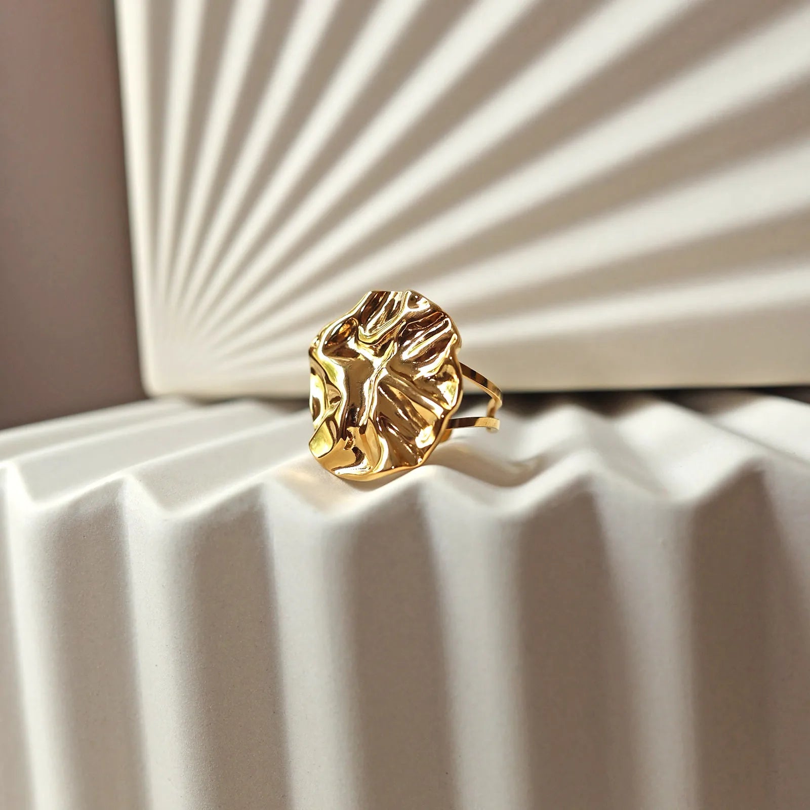 Bague ajustable Je me dévoile en acier doré, à motif sculpté, posée sur un fond beige texturé aux reflets doux.