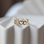 Bague "Unité" réglable en acier doré, composée de cœurs entrelacés, posé sur un support beige texturé.