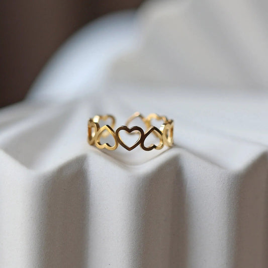 Bague "Unité" réglable en acier doré, composée de cœurs entrelacés, posé sur un support beige texturé.