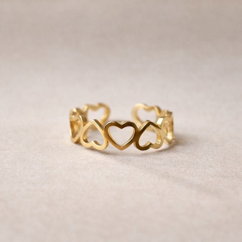 Bague "Unité" réglable en acier doré, composée de cœurs entrelacés, posé sur un support beige texturé.