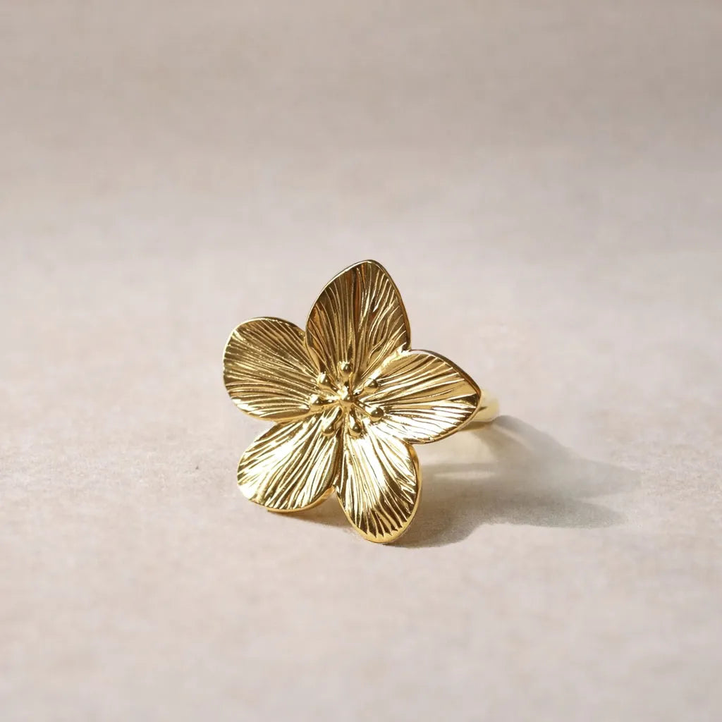 Bague réglable dorée en acier inoxydable, design fleur, vue de face, posée sur un support beige texturé.