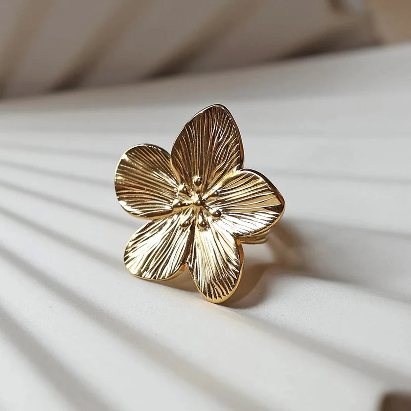 Bague réglable dorée en acier inoxydable, design fleur, vue rapprochée de face, posée sur un support beige texturé.