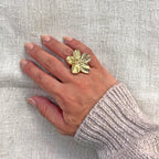 Bague réglable dorée en acier inoxydable, design fleur , portée sur une main féminine avec un pull en maille claire, posée sur un tissu beige texturé.