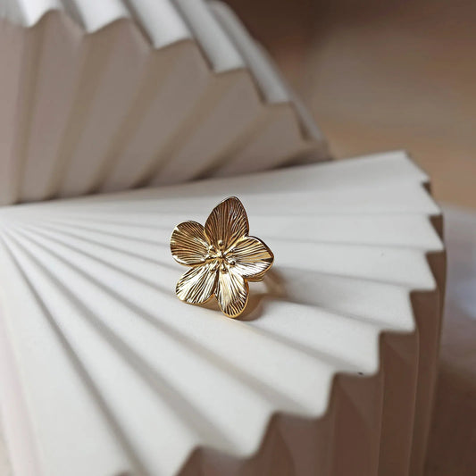 Bague réglable dorée en acier inoxydable, design fleur, vue de face, posée sur un support beige texturé.
