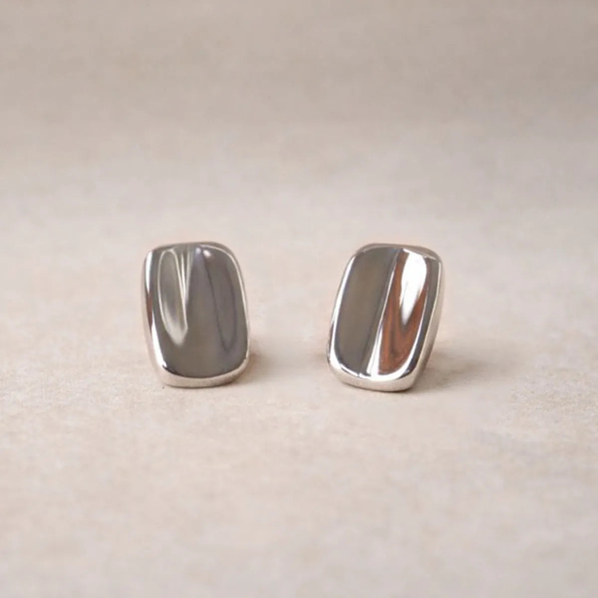 Vue rapprochée des boucles d’oreilles Généreuse minimalistes rectangulaires  en acier inoxydable argenté, posées sur un fond beige texturé.