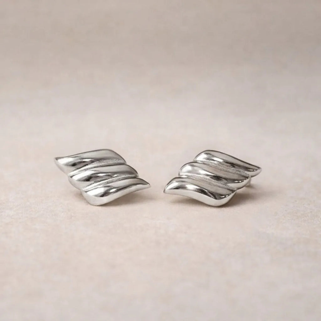 Boucles d'oreilles Envolée, ailes argentées en acier inoxydable,  sur un fond texturé beige clair.