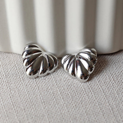 Boucles d'oreilles "Coeur audacieux", en acier inoxydable argenté, au design sculpté en forme de coeur, capturées en gros plan sur fond texturé beige.