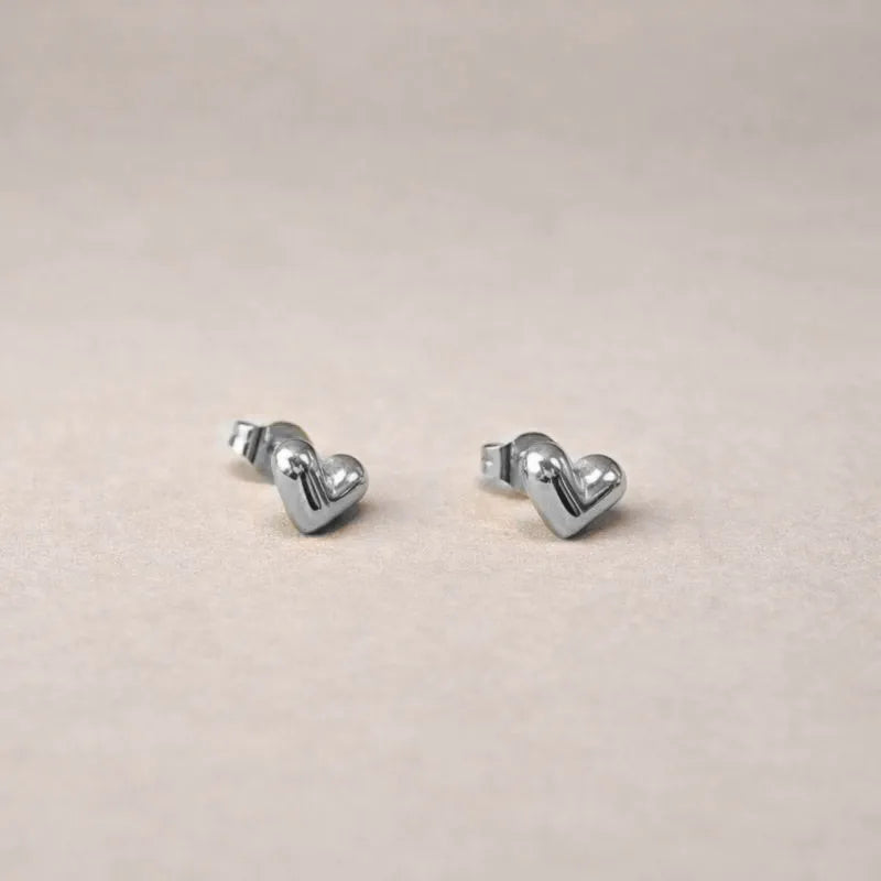 Zoom sur les boucles d'oreilles "Tendre discrétion" de Visée, en forme de cœur en acier argenté, posées sur un fond beige texturé.