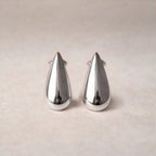 Boucles d'oreilles Pure en  acier argenté, en forme de goutte, posées sur un fond  beige texturé.