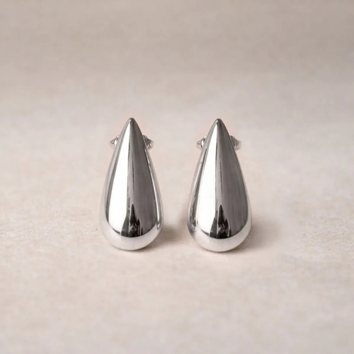 Boucles d'oreilles Pure en  acier argenté, en forme de goutte, posées sur un fond  beige texturé.
