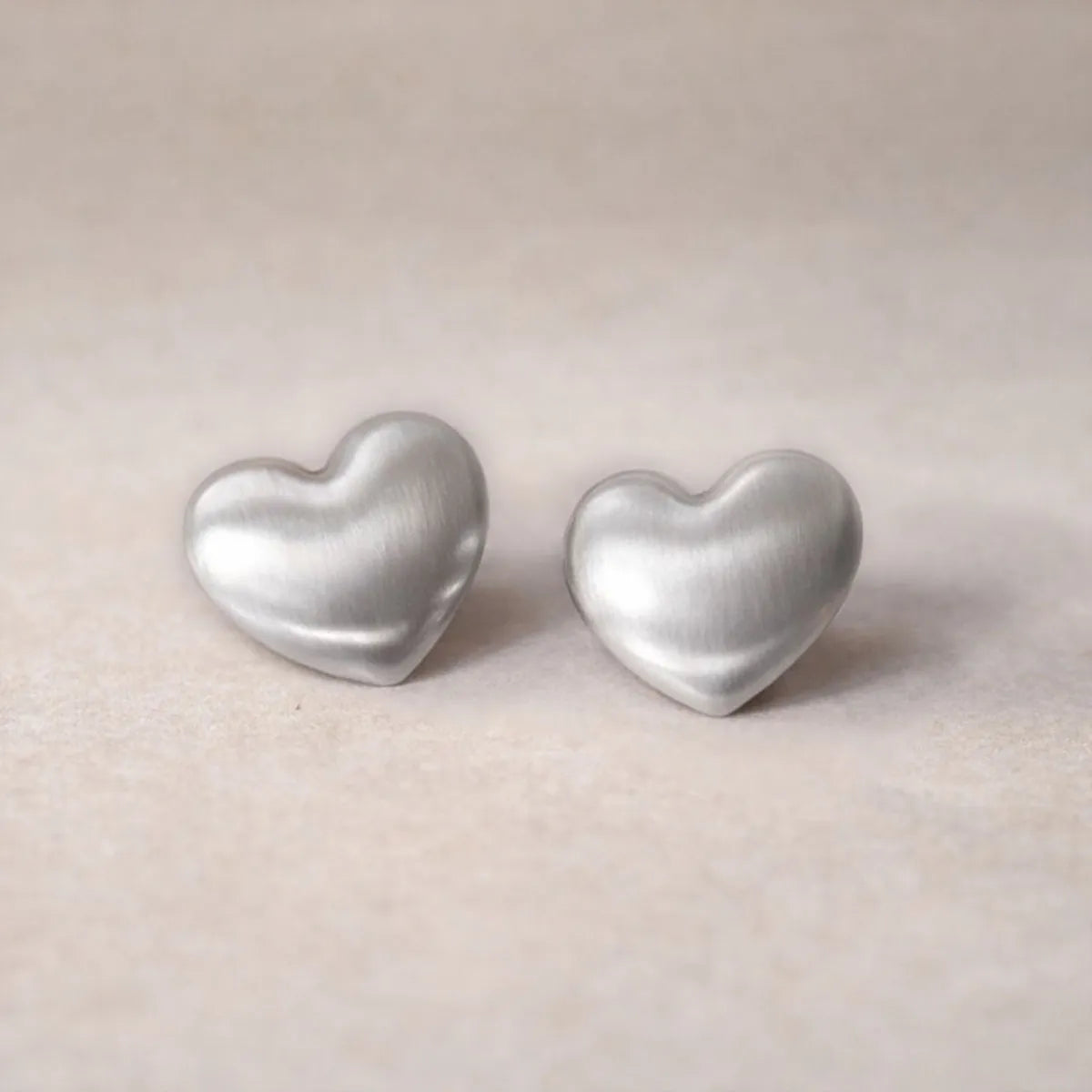 Zoom sur les boucles d'oreilles "S'aimer", en maxi cœurs argentés mat, en acier Inoxydable, posées sur un fond beige texturé.