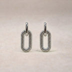 Boucles d'oreilles "Modulable" en acier argenté, au design modulable, photographiées sur un tissu beige texturé.