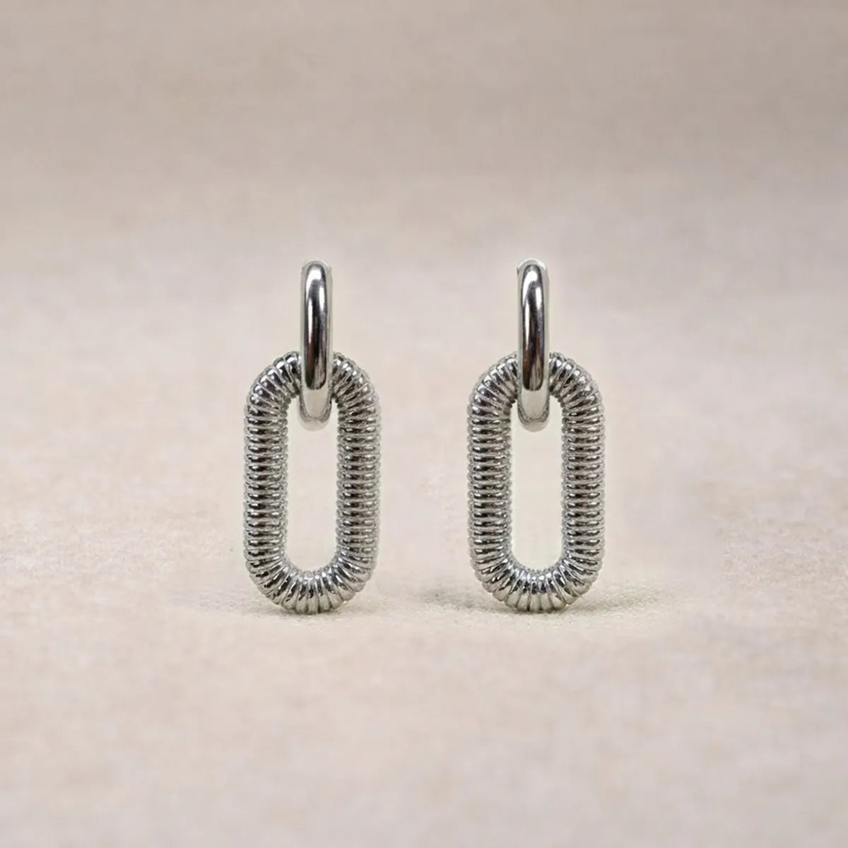 Boucles d'oreilles "Modulable" en acier argenté, au design modulable, photographiées sur un tissu beige texturé.